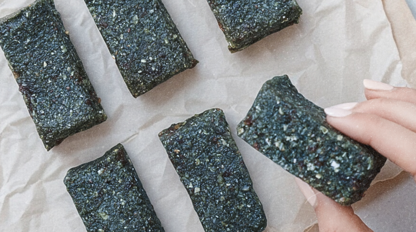 Hand holding a superfood-packed Oatmeal Date Bar made with Elyxar—dark, chewy bars arranged on parchment paper for a nutritious, no-bake snack.