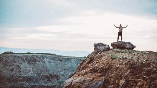 Person standing with arms raised on a rocky summit — symbolizing the natural strength, clarity, and vitality fueled by Elyxar from Apotheka Superfoods.